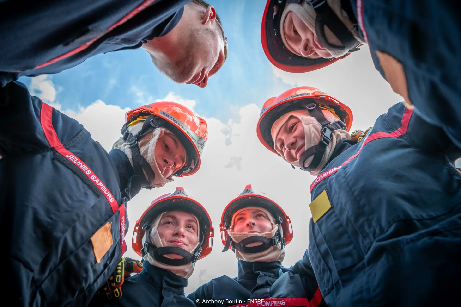 Vue d'en bas d'un groupe de cinq jeunes sapeurs-pompiers, casqués et vêtus de leur uniforme, formant un cercle et regardant vers le haut, sous un ciel bleu avec quelques nuages.