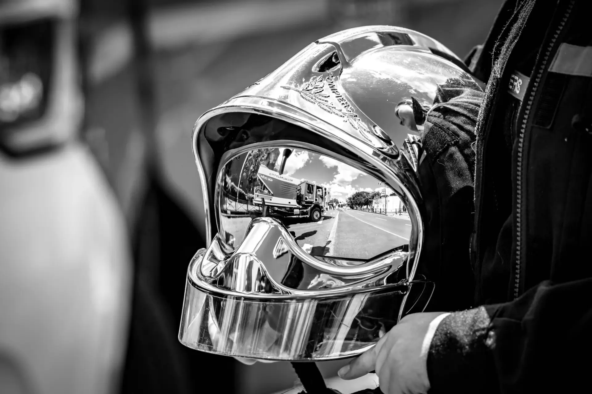 Une photo en noir et blanc d'un casque de pompier brillant tenu par un pompier. Le casque reflète une scène de rue avec un camion de pompiers et des arbres.