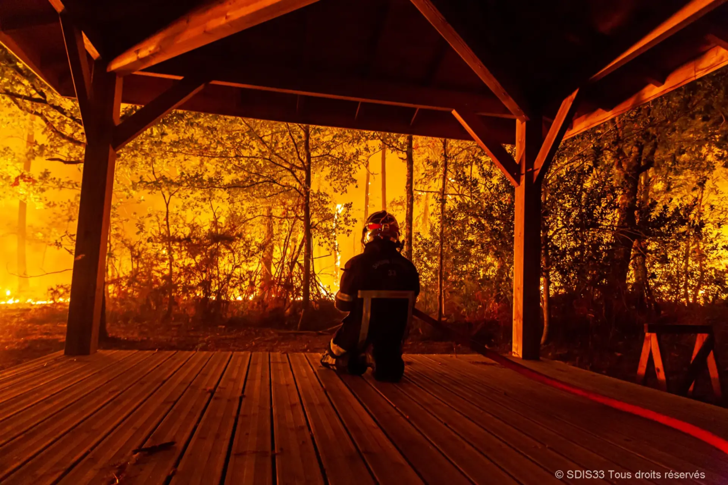 Un pompier est agenouillé sous un auvent en bois, observant un incendie de forêt intense d'une lueur orange et rouge.