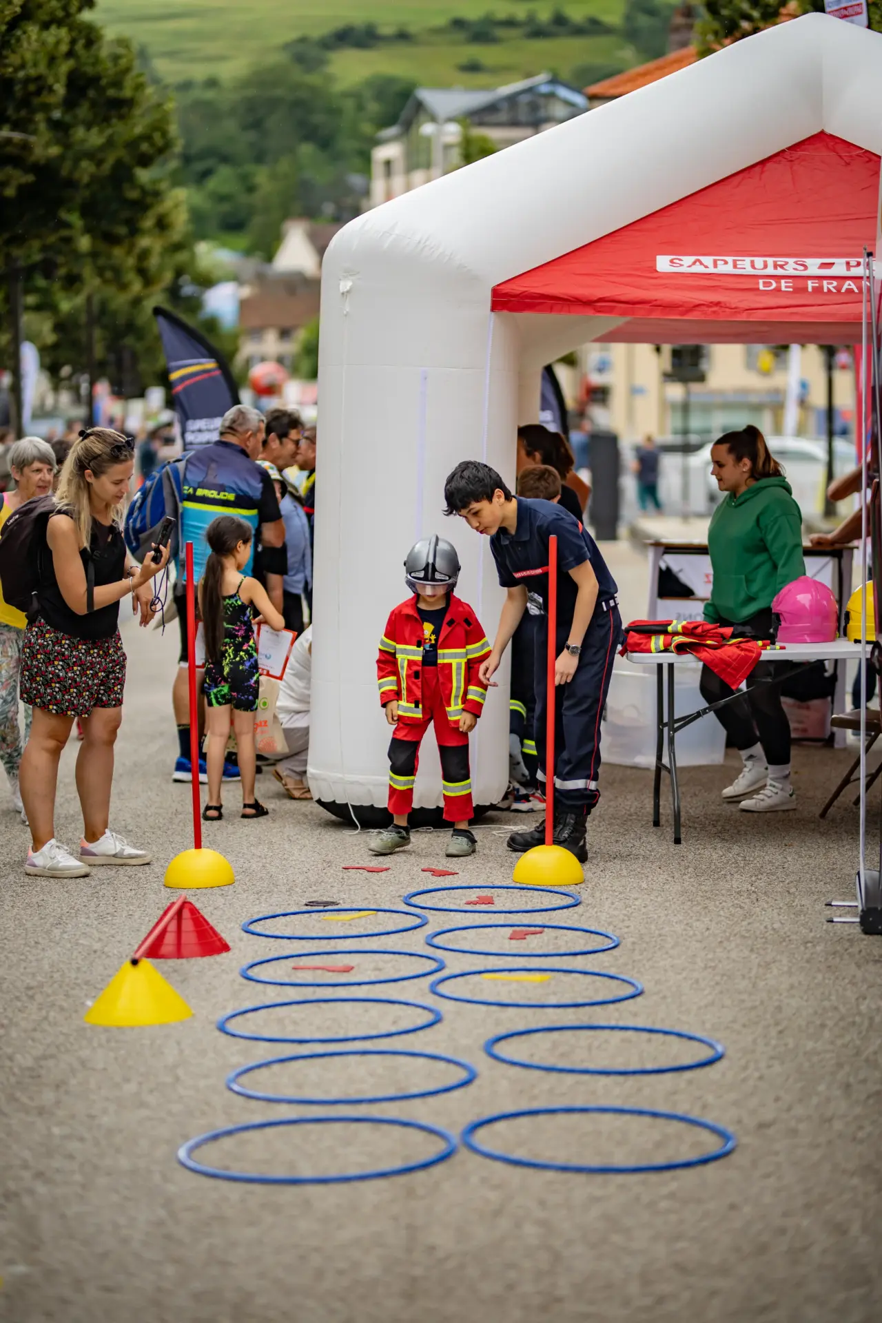Un pompier aide un jeune enfant déguisé en pompier à traverser un parcours d'agilité fait de cerceaux bleus et de cônes jaunes, sous un stand rouge et blanc des "SAPEURS-POMPIERS DE FRANCE".